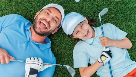 father and son laying down on grass smiling holding golf clubs