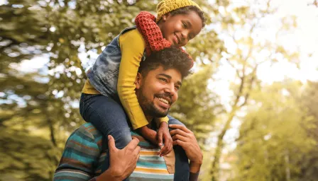 A girl riding on her dads shoulders outside with sun shining
