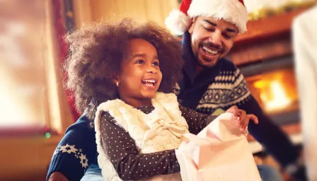 Smiling man in Santa hat watching smiling child open a present